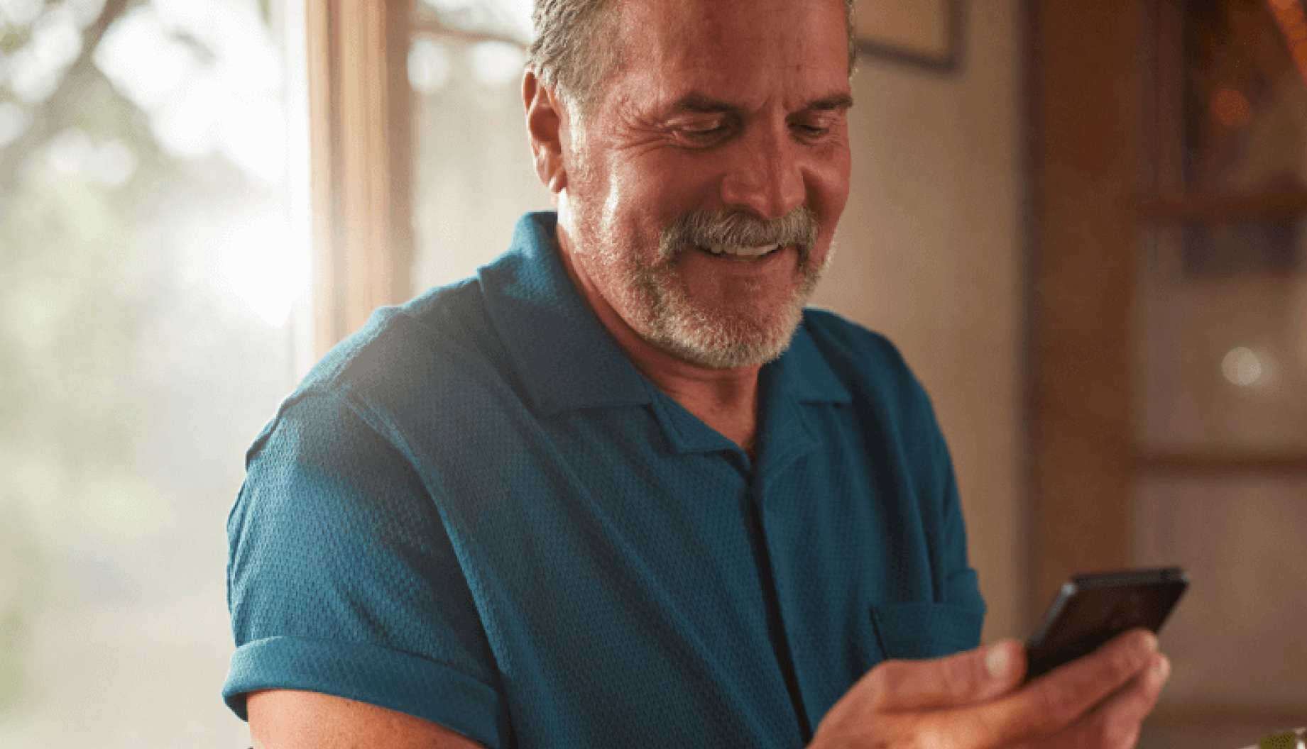An Eversense user in a blue shirt smiles while looking at a phone, standing near a bright window indoors.